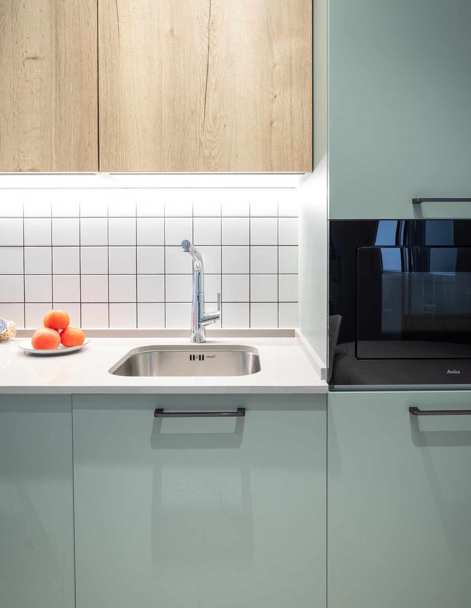 Fregadero de cocina con encimera blanca, azulejos cuadrados y muebles verde menta.